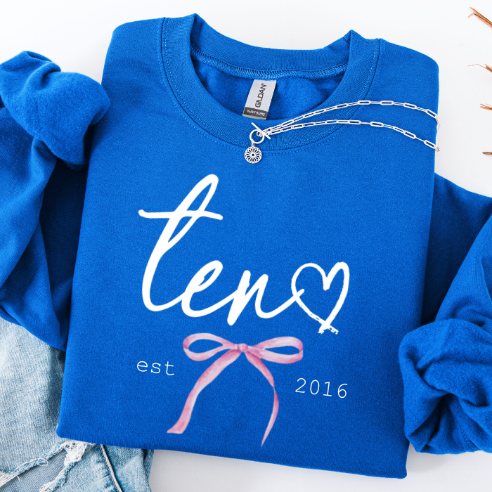 A folded royal blue sweatshirt with rolled sleeves, featuring a white script 'ten' with a heart, 'est 2016' text, and a pink bow graphic. A silver chain necklace is draped across the chest, and light blue denim jeans are partially visible at the bottom left.
