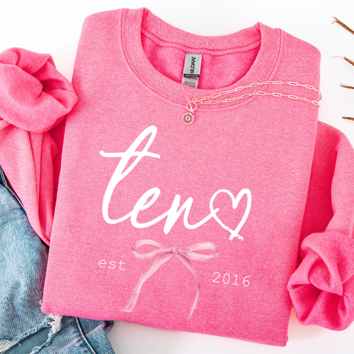 A folded pink sweatshirt with rolled sleeves, featuring a white script graphic that reads 'ten' with a heart, plus 'est. 2016' and a bow illustration.