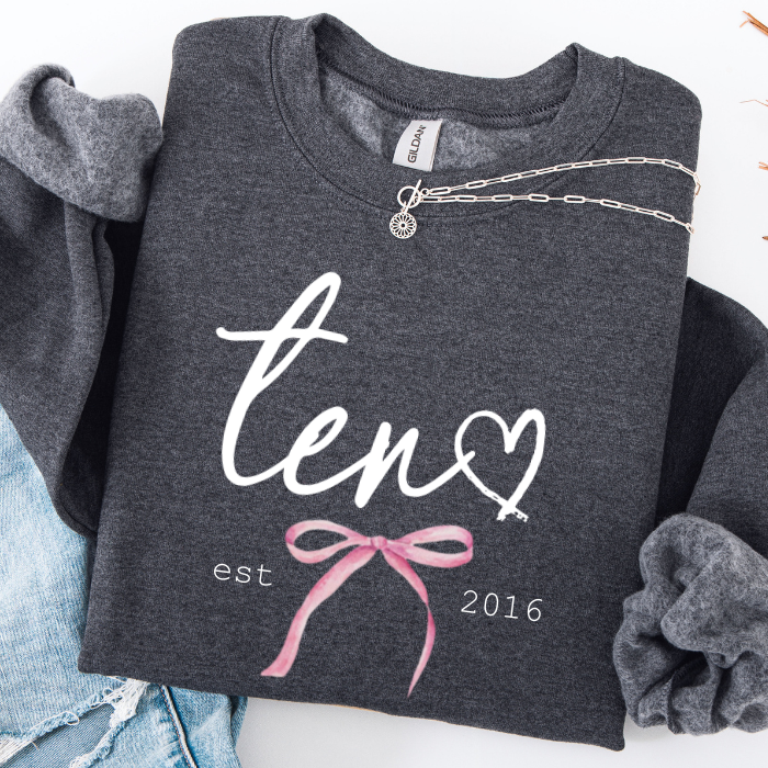 A dark heather gray sweatshirt laid flat on a white background, styled with rolled sleeves, a silver chain necklace, and light blue denim. The sweatshirt has a white script graphic reading 'ten' with a heart, plus 'est 2016' and a pink bow illustration.