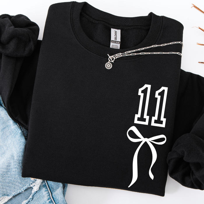 A folded black long-sleeve sweatshirt with a white graphic print of the number 11 and a bow on the front, styled with a silver chain necklace and light blue denim shorts on a white background.