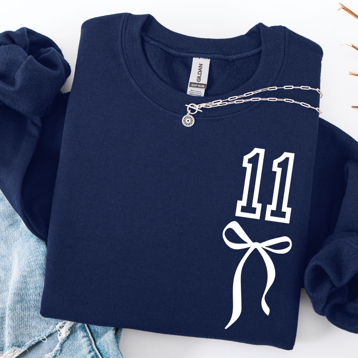 A navy blue sweatshirt with a white graphic print of the number 11 and a ribbon bow on the front, styled flat with a silver chain necklace and light blue denim jeans.