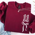 A folded burgundy crewneck sweatshirt with a white graphic print of the number 11 and a bow on the front, styled with rolled sleeves and a silver chain necklace on a white background.
