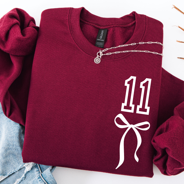A folded burgundy crewneck sweatshirt with a white graphic print of the number 11 and a bow on the front, styled with rolled sleeves and a silver chain necklace on a white background.