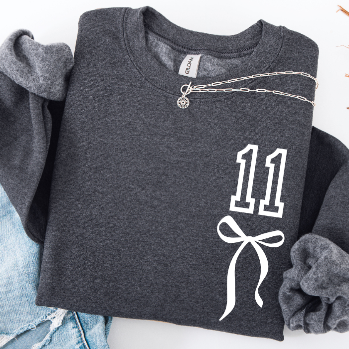 A dark gray crewneck sweatshirt with a white graphic print of the number 11 and a bow on the front, styled in a flat lay with a silver chain necklace, blue jeans, and a gray scrunchie on a white background.