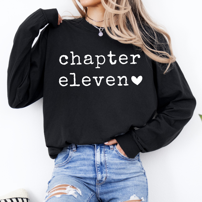 A woman wearing a black long-sleeve sweatshirt with 'chapter eleven' printed in white typewriter-style text and a small white heart, paired with distressed blue jeans.