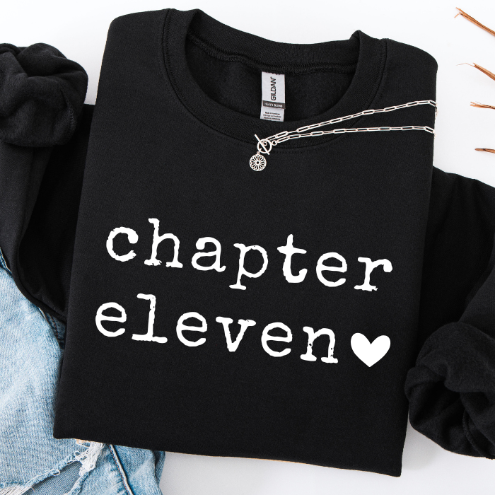 A black crewneck sweatshirt with 'chapter eleven' and a heart printed in white on the front, styled flat-lay with a silver necklace and light blue denim.
