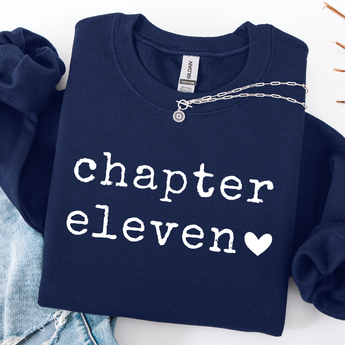 A navy blue sweatshirt with 'chapter eleven' and a heart printed in white on the front, styled flat-lay with a silver chain necklace and light blue denim.
