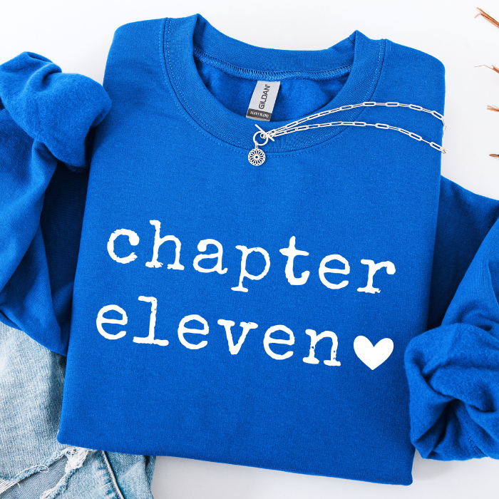 A folded royal blue sweatshirt with rolled sleeves, featuring the words 'chapter eleven' in white lowercase letters and a white heart on the front. A silver chain necklace is draped across the chest, and light-wash denim jeans are partially visible at the bottom left.