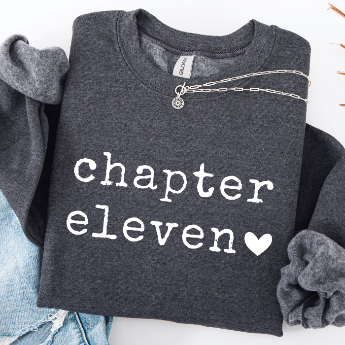 A dark heather gray sweatshirt with 'chapter eleven' printed in white typewriter-style text and a white heart, styled flat-lay with rolled sleeves, a silver chain necklace, and light blue jeans on a white background.