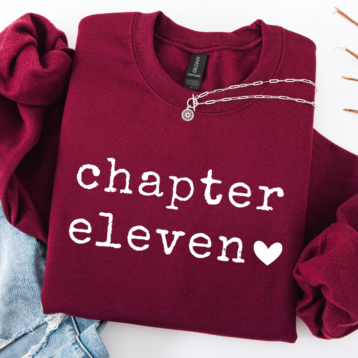 A folded burgundy sweatshirt with 'chapter eleven' and a heart printed in white on the front, styled with a silver chain necklace and light blue jeans on a white background.