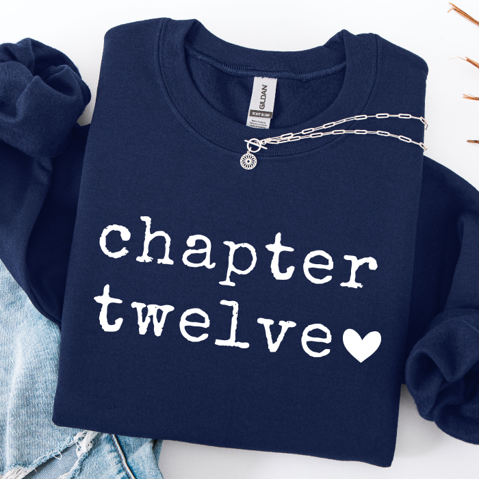 A navy blue sweatshirt with 'chapter twelve' printed in white typewriter-style text and a white heart, styled flat-lay with light blue jeans and a silver chain necklace on a white background.