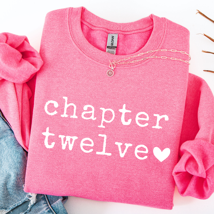 A folded pink long-sleeve sweatshirt with rolled sleeves, featuring the text 'chapter twelve' in white typewriter-style lettering and a white heart. A silver chain necklace is draped across the chest.