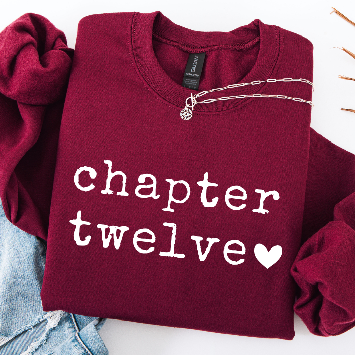 A folded burgundy sweatshirt with 'chapter twelve' printed in white across the front, styled with a silver chain necklace and light blue denim on a white background.