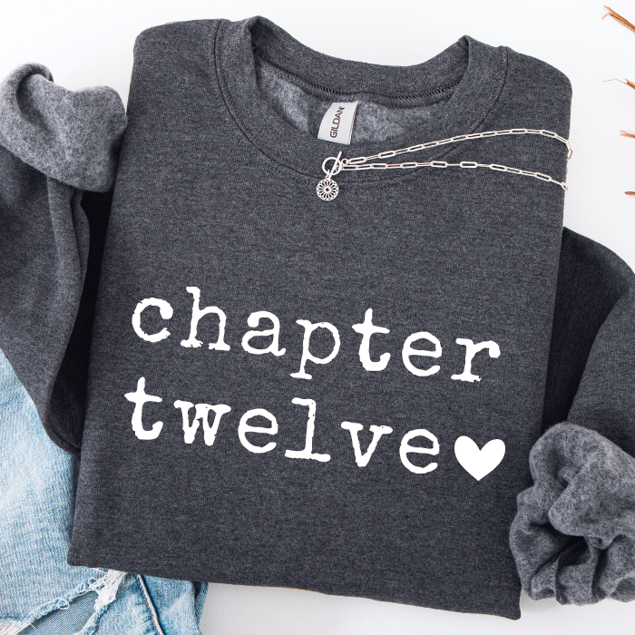 A dark heather gray sweatshirt with 'chapter twelve' printed in white typewriter-style text and a small white heart, styled flat-lay with rolled sleeves, a silver chain necklace, and light blue jeans on a white background.