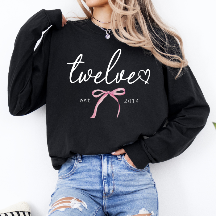 A woman wearing a black long-sleeve sweatshirt with a graphic that reads 'twelve' in white script, a pink bow illustration, and 'est 2014' in small text, paired with distressed blue jeans.