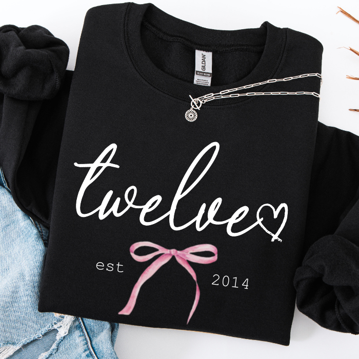 A folded black sweatshirt with white script text reading 'twelve' and a pink ribbon graphic, styled on a white surface with denim and a silver necklace.