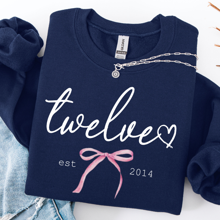 A navy blue sweatshirt with 'twelve' in white script, a small white heart, 'est 2014' in white text, and a pink ribbon graphic on the front. The sweatshirt is styled flat with a silver chain necklace draped across the chest and light blue denim jeans partially visible at the bottom left.