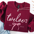 A folded burgundy sweatshirt with white script text reading 'twelve' and a pink bow graphic, styled on a white background with rolled sleeves and a silver necklace draped across the collar.