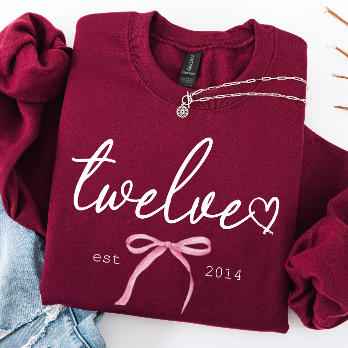 A folded burgundy sweatshirt with white script text reading 'twelve' and a pink bow graphic, styled on a white background with rolled sleeves and a silver necklace draped across the collar.