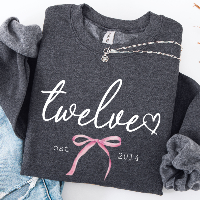 A dark heather gray sweatshirt laid flat on a white background, featuring the word 'twelve' in white script with a heart, 'est 2014' in white text, and a pink ribbon graphic. A silver chain necklace is draped across the chest.