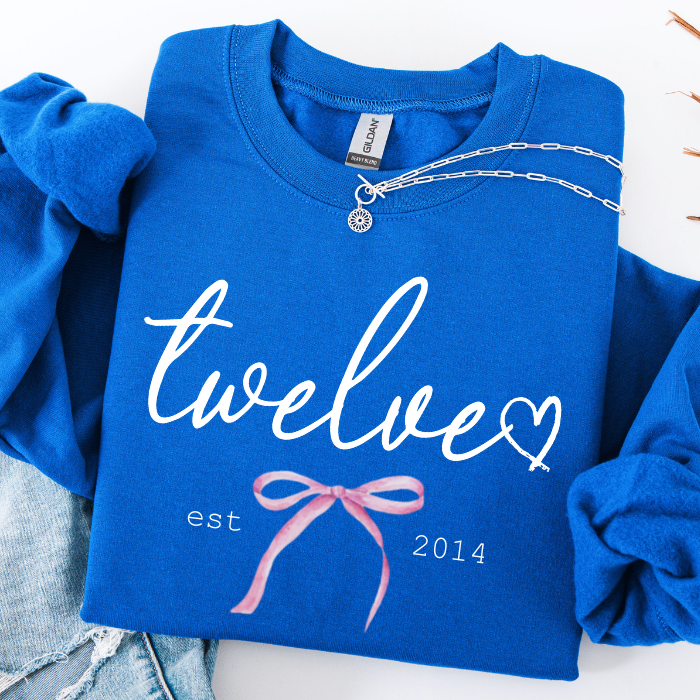 A folded royal blue sweatshirt with rolled sleeves, featuring white script text that reads 'twelve' with a heart, 'est 2014', and a pink ribbon graphic. A silver necklace is draped across the collar, and light blue jeans are partially visible at the bottom left.