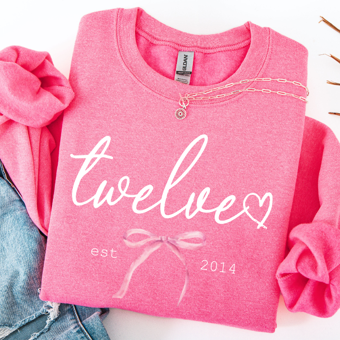 A folded pink sweatshirt with rolled sleeves, featuring the word 'twelve' in white script with a heart, 'est. 2014' in small text, and a pale pink bow graphic.