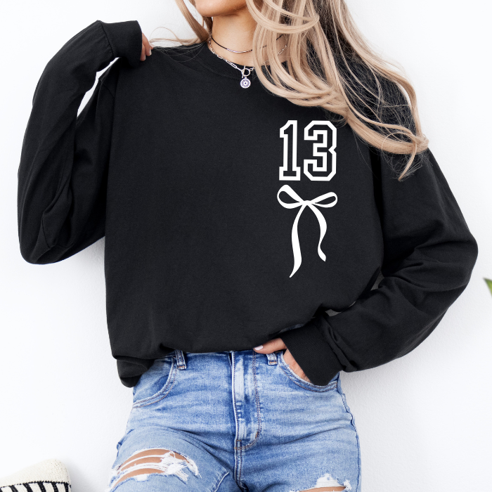 A woman wearing a black long-sleeve sweatshirt with a white graphic print of the number 13 and a bow on the front, paired with distressed blue jeans.