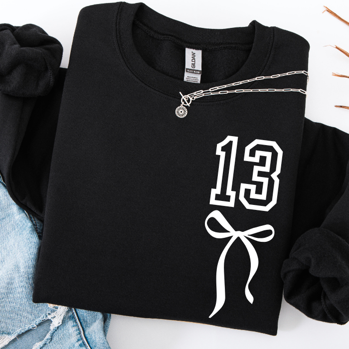 A black crewneck sweatshirt with a white graphic print of the number 13 and a bow, styled flat-lay with a silver necklace and light blue denim.