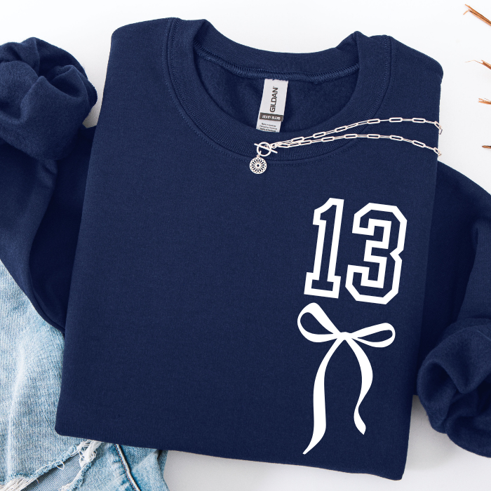A navy blue sweatshirt with a white '13' and bow graphic on the front, styled flat-lay with a silver chain necklace draped across the chest and light blue denim at the side.