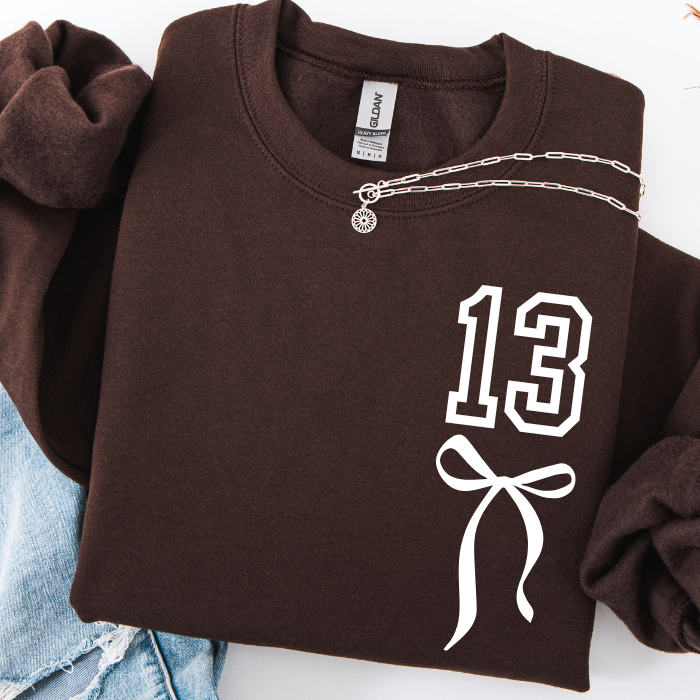 A folded dark brown sweatshirt with a white outlined '13' and bow graphic on the front, styled with a silver chain necklace and light blue jeans on a white background.