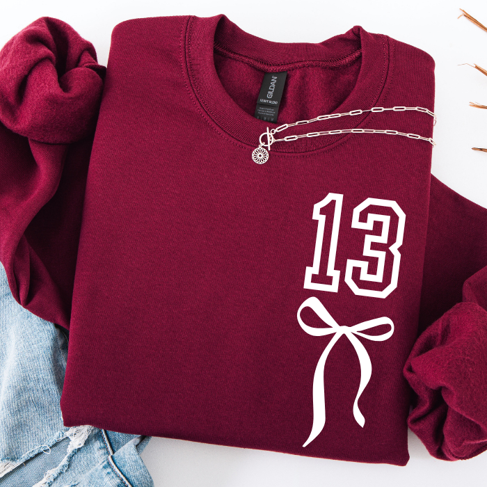 A folded burgundy sweatshirt with a white outlined '13' and bow graphic on the front, styled with rolled sleeves, a silver chain necklace, and light blue denim on a white background.