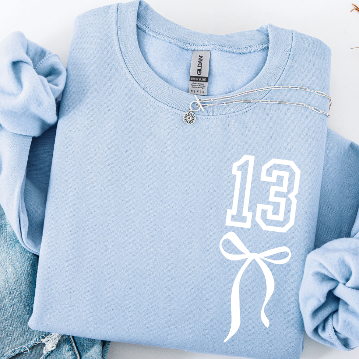 A folded light blue Gildan t-shirt with a white graphic print of the number 13 and a bow, styled with rolled sleeves and a silver necklace on a white background.