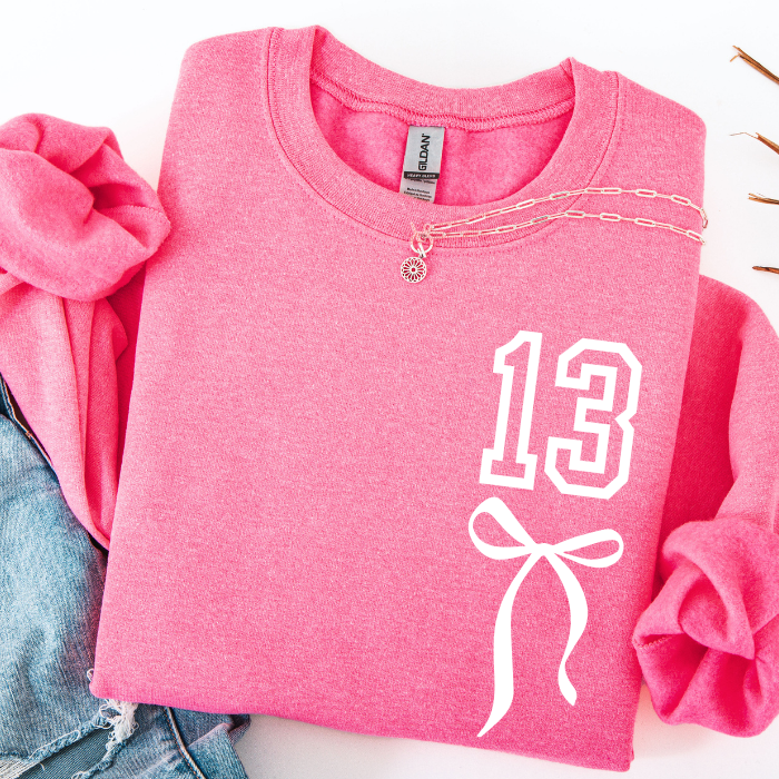 A folded pink sweatshirt with rolled sleeves, featuring a white graphic of the number 13 and a bow on the front. A gold chain necklace is draped across the chest.