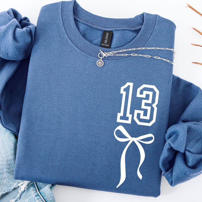 A folded blue sweatshirt with rolled sleeves, featuring a white outlined '13' and bow graphic on the front. A silver chain necklace is draped across the chest, and the sweatshirt is styled with light denim jeans on a white background.