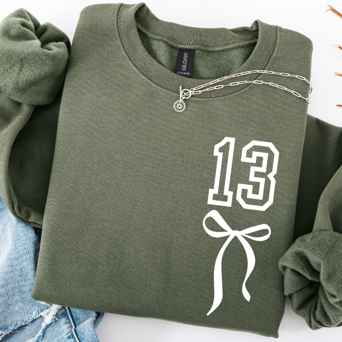 An olive green crewneck sweatshirt with rolled sleeves, featuring a white graphic print of the number '13' and a bow on the front. A silver chain necklace is draped across the collar.