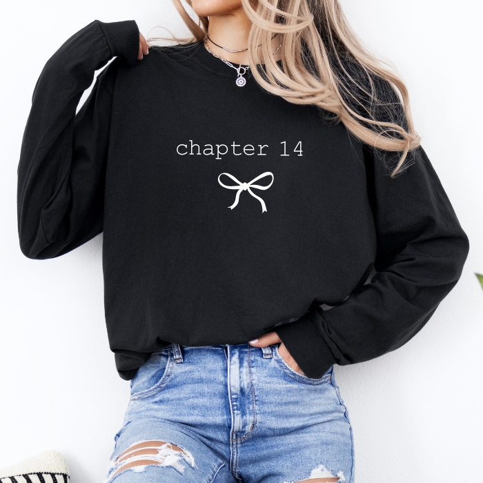 A woman wearing a black long-sleeve sweatshirt with 'chapter 14' and a bow graphic printed in white on the chest, paired with distressed blue jeans.