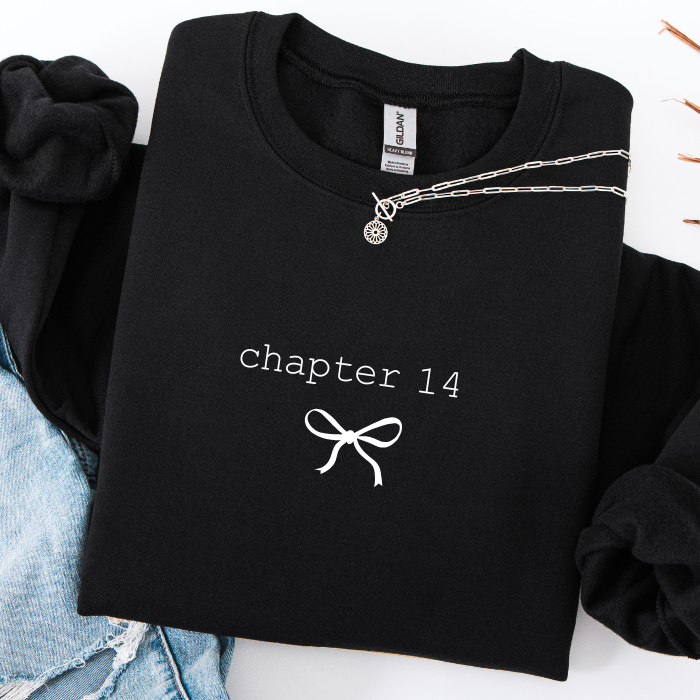 A folded black sweatshirt with 'chapter 14' and a bow graphic printed in white on the front, styled with a silver chain necklace and light blue denim on a white background.