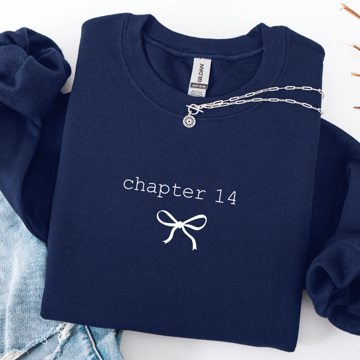 A navy blue sweatshirt with 'chapter 14' and a bow graphic printed in white on the front, styled flat-lay with light blue denim and a silver chain necklace.