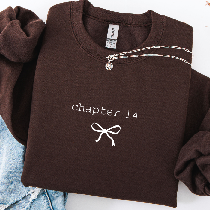 A folded dark brown sweatshirt with 'chapter 14' and a bow graphic printed in white on the front, styled with rolled sleeves and a silver chain necklace draped across the neckline. A denim fabric is visible at the lower left.