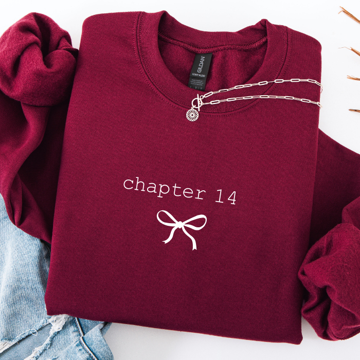 A folded burgundy sweatshirt with 'chapter 14' and a bow graphic printed on the front, styled with a silver chain necklace and light blue denim on a white background.