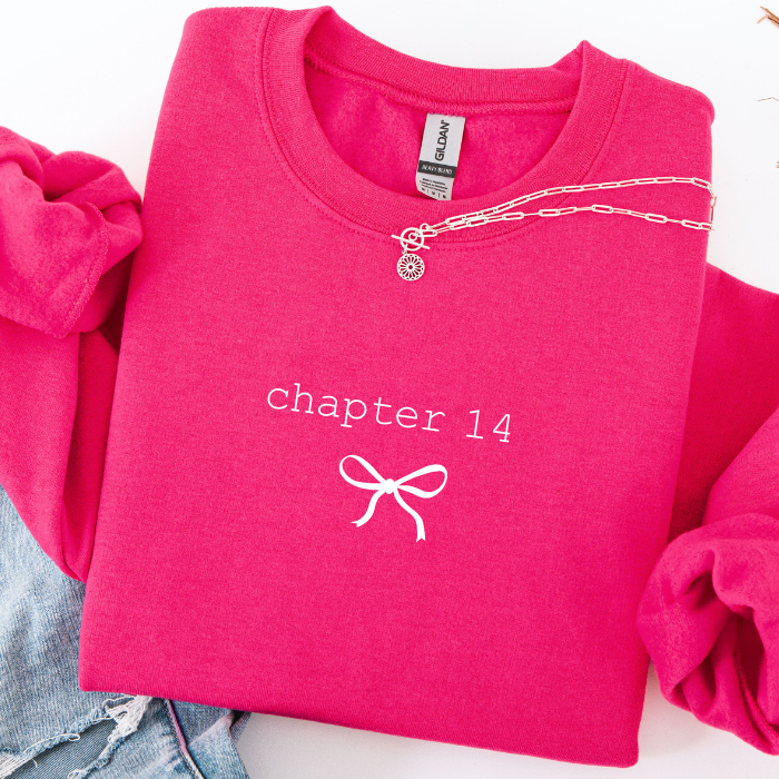 A folded hot pink sweatshirt with 'chapter 14' and a bow graphic printed in white on the front, styled with rolled sleeves and denim on a white background.
