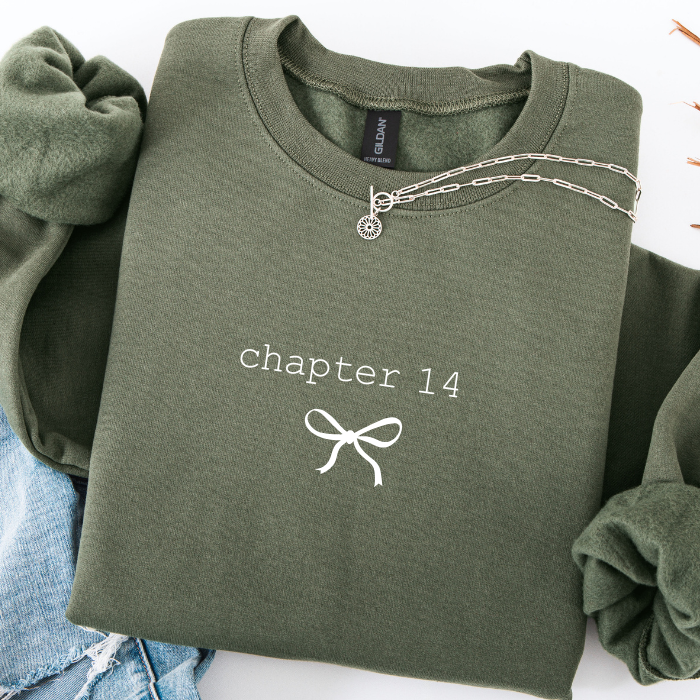 An olive green crewneck sweatshirt with 'chapter 14' and a bow graphic printed on the chest, styled flat with a silver chain necklace and blue jeans.