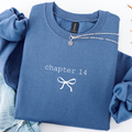 A folded blue sweatshirt with 'chapter 14' and a bow graphic printed in white on the front, styled with rolled sleeves and a silver chain necklace on a white background.