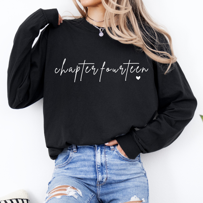 A woman wearing a black long-sleeve sweatshirt with 'chapter fourteen' printed in white script across the chest, paired with distressed blue jeans.