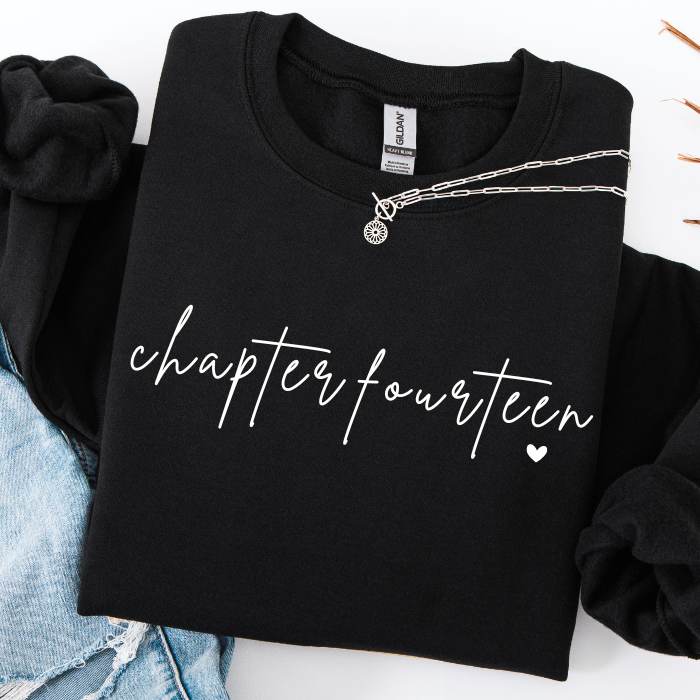 A folded black long-sleeve sweatshirt with 'chapter fourteen' printed in white script across the front, styled with a silver chain necklace and light blue denim on a white background.