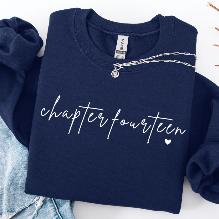 A navy blue sweatshirt with 'chapter fourteen' and a small heart printed in white across the front, styled flat-lay with light blue denim and a silver chain necklace on a white background.