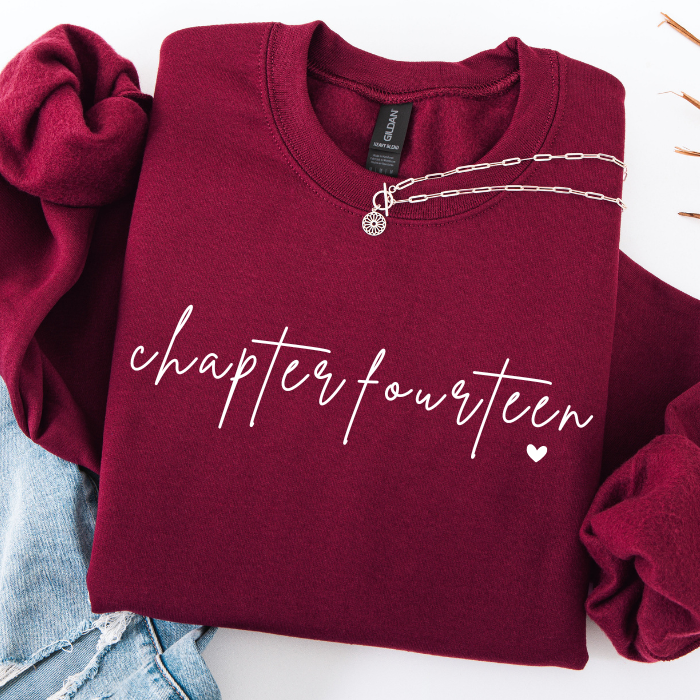 A folded burgundy sweatshirt with 'chapter fourteen' printed in white script across the front, styled with a silver chain necklace and light blue denim on a white background.