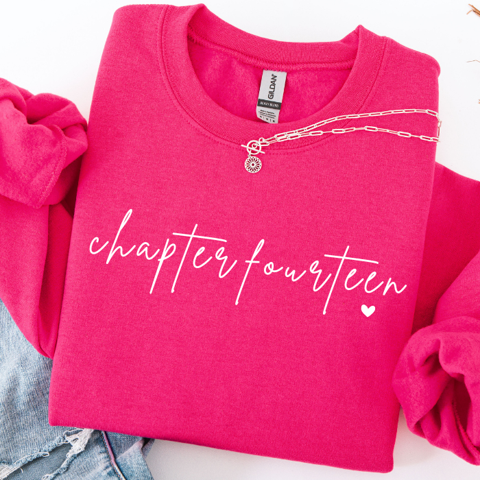 A folded hot pink sweatshirt with the words 'chapter fourteen' and a small heart printed in white across the front. A silver chain necklace is draped across the chest, and part of a pair of blue jeans is visible at the lower left.