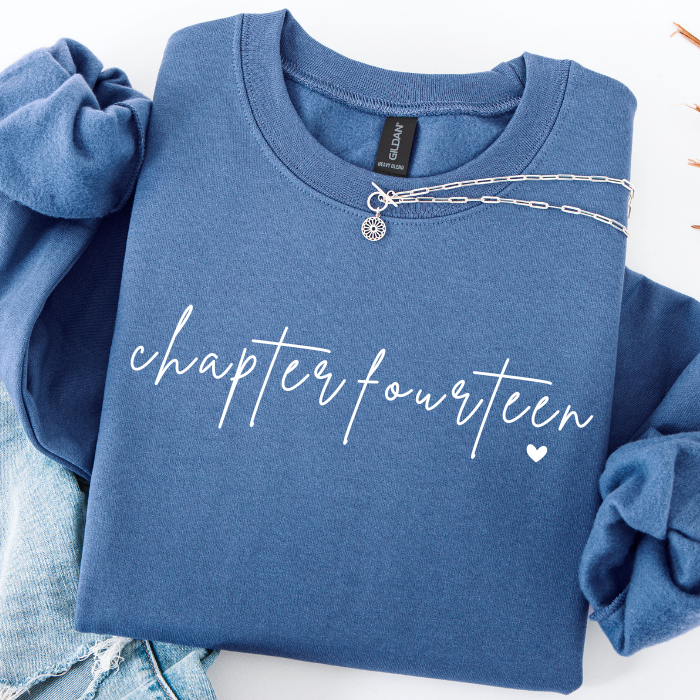 A folded blue sweatshirt with rolled sleeves, featuring the words 'chapter fourteen' in white script across the front, styled with a silver chain necklace and light-wash denim on a white background.