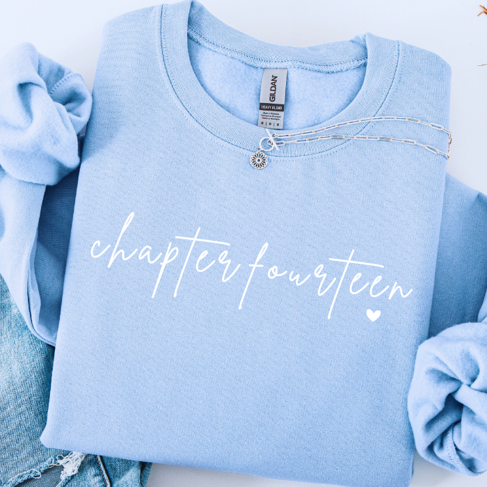 A folded light blue crewneck sweatshirt with 'chapter fourteen' printed in white script across the front, styled with rolled sleeves and a silver necklace on a white background.